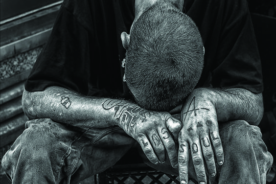 Black and white image of a man wearing jeans and black t-shirt, with his head on his hands, which have several tattoos including the words 'LOST SOUL'