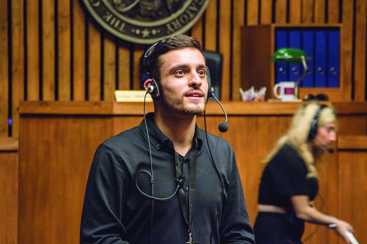 A student stage manager stands on a wooden panelled set of a courtroom, speaking into a headset