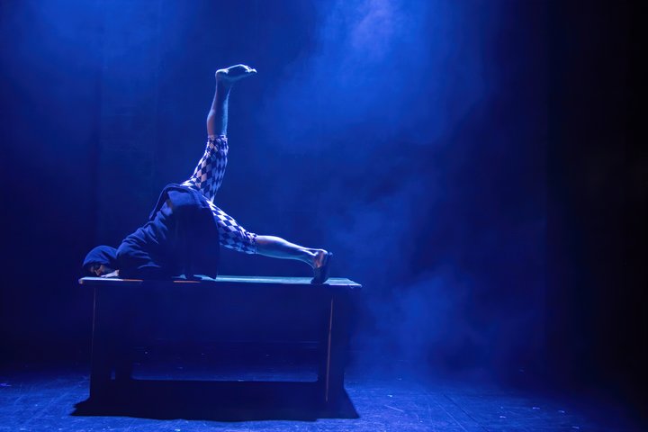 An actor performs on a table in a dark dressing gown and a black and white clown-esque trouser. They perform in dramatic blue lighting.