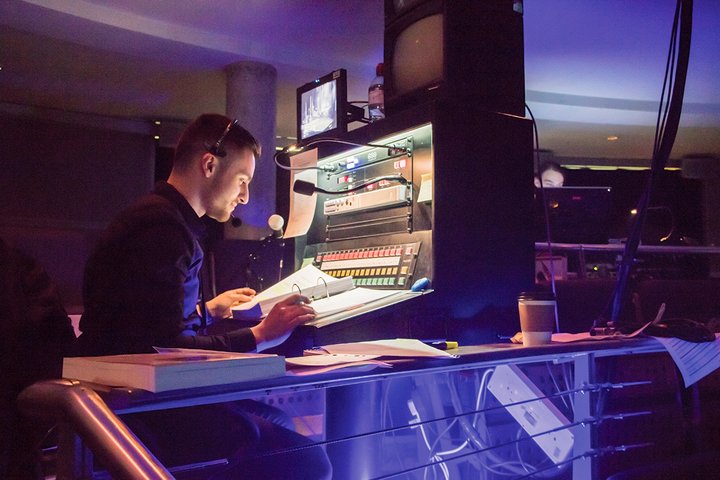 A stage manager sits at the prompt desk wearing a headset and lit in pink and blue hues.