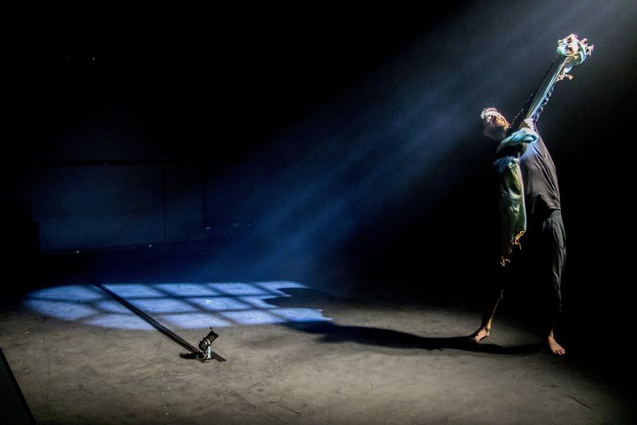 A man stands on stage, he is dimly lit. A projection of a window is next to him.