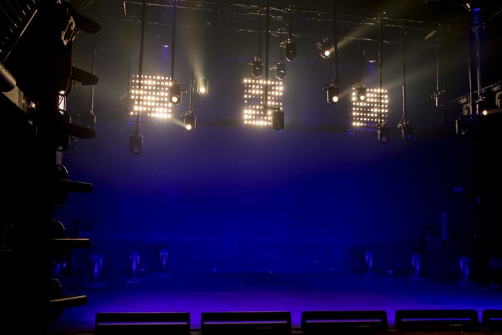 The Jerwood Vanbrugh Theatre is in an indigo blue light wash. Three large lights illuminate a silhouette of more theatre lights hanging from the rig.