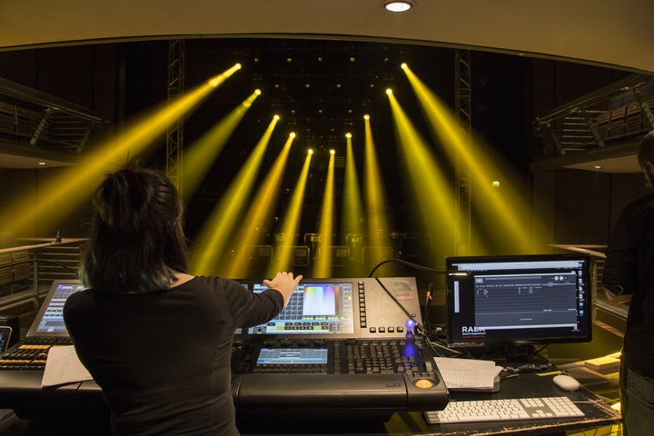 A view from the back of a theatre, behind a student who is programming a lighting desk. Yellow lights fill the stage and auditorium.