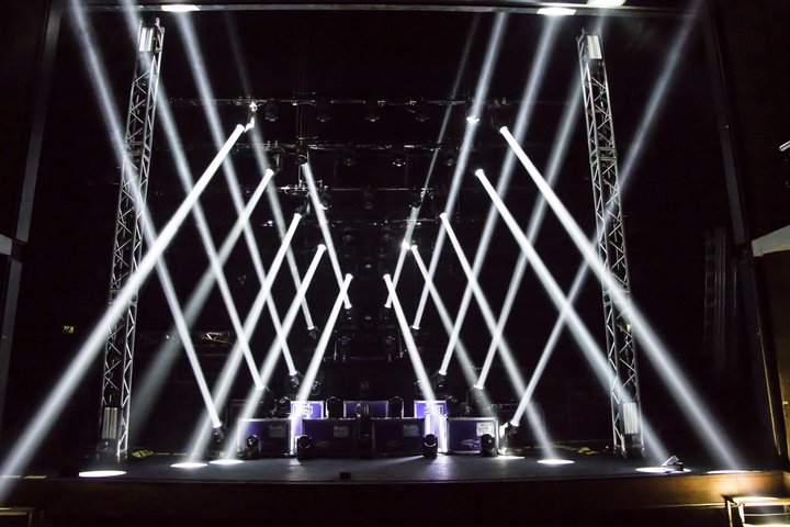 A wide view of the expanse of the Vanburgh Theatre stage filled with lighting equipment. Striking white lights criss cross across the stage