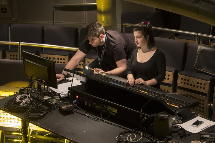 Two students programme the lighting desk in theatre Vanbrugh Theatre at RADA as yellow stage lights reflect from the stage onto the seats around them.