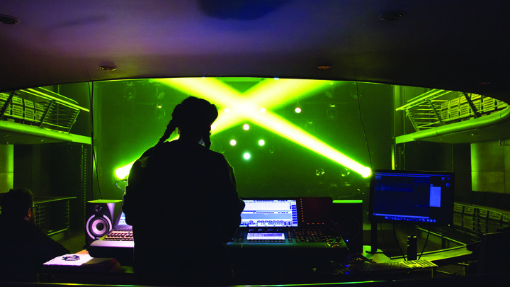 Viewed from behind a theatre lighting desk, a woman programmes a lighting desk as lime green and yellow spotlights shine across the stage