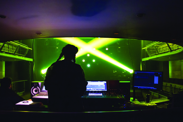 Viewed from behind a theatre lighting desk, a woman programmes a lighting desk as lime green and yellow spotlights shine across the stage