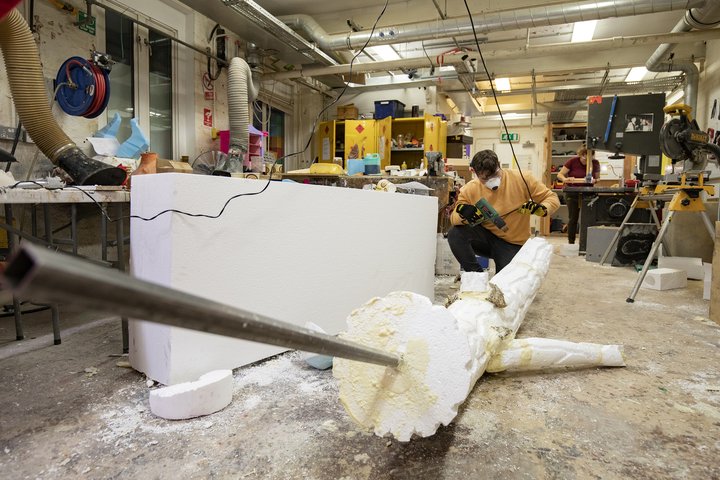 In a workshop, a student kneels by a polystyrene model of a tree branch, which extends out towards the camera, using a drill to shape a bark pattern.