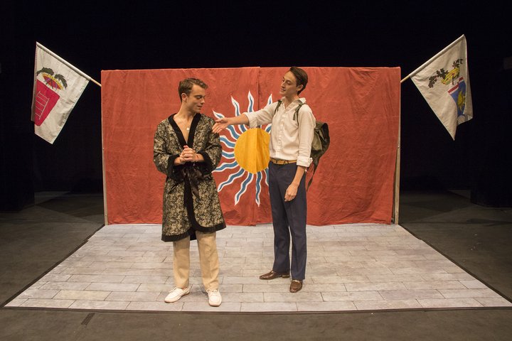 Two actors stand on stage in front of an orange cloth backdrop with flags and sun insignia; one offers the other his hand to shake