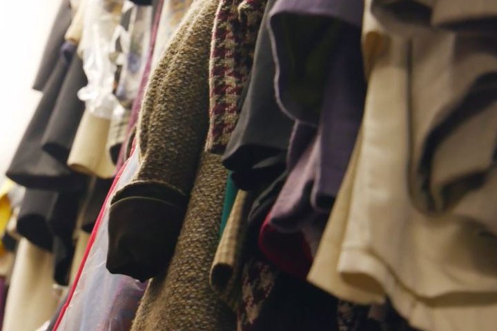 We are looking up into a selection of costumes of varying textures and colours, with a tweed coat in the middle in sharpest focus, hanging on a rail.