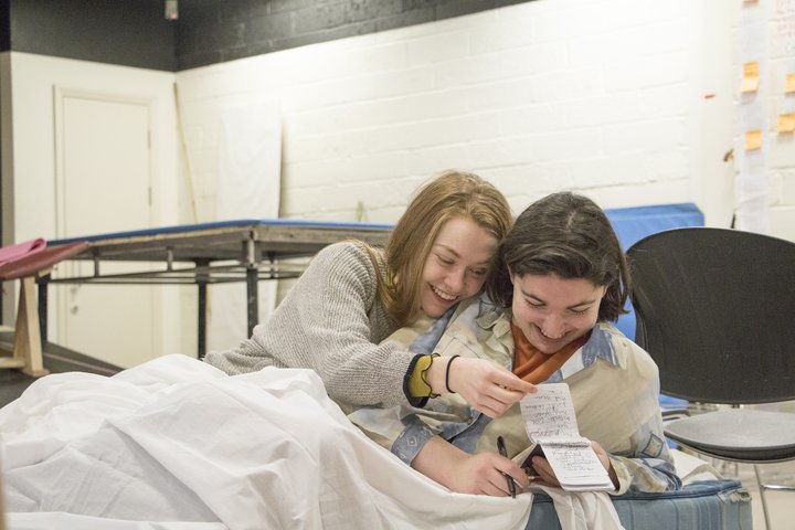 Two female acting students lie close in bed, laughing as they flick through a small notebook filled with scrawled handwriting.