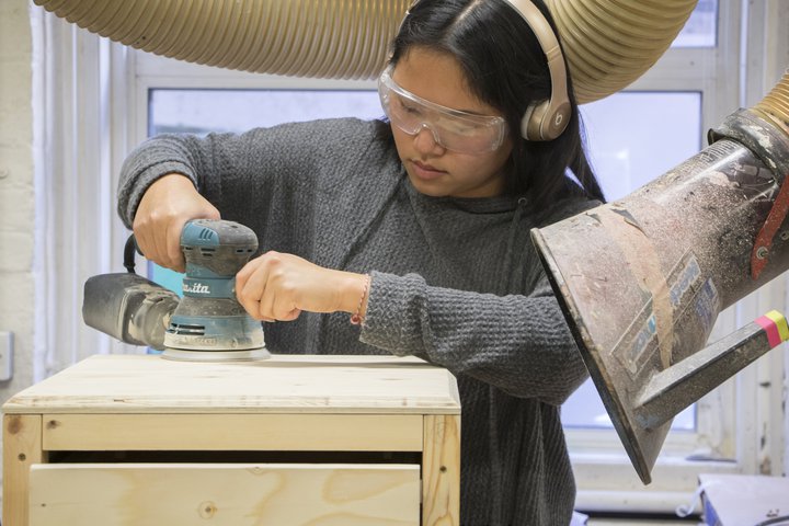 © A female student wearing safety glasses uses a sanding machine on a piece of wooden furniture in a workshop at RADA